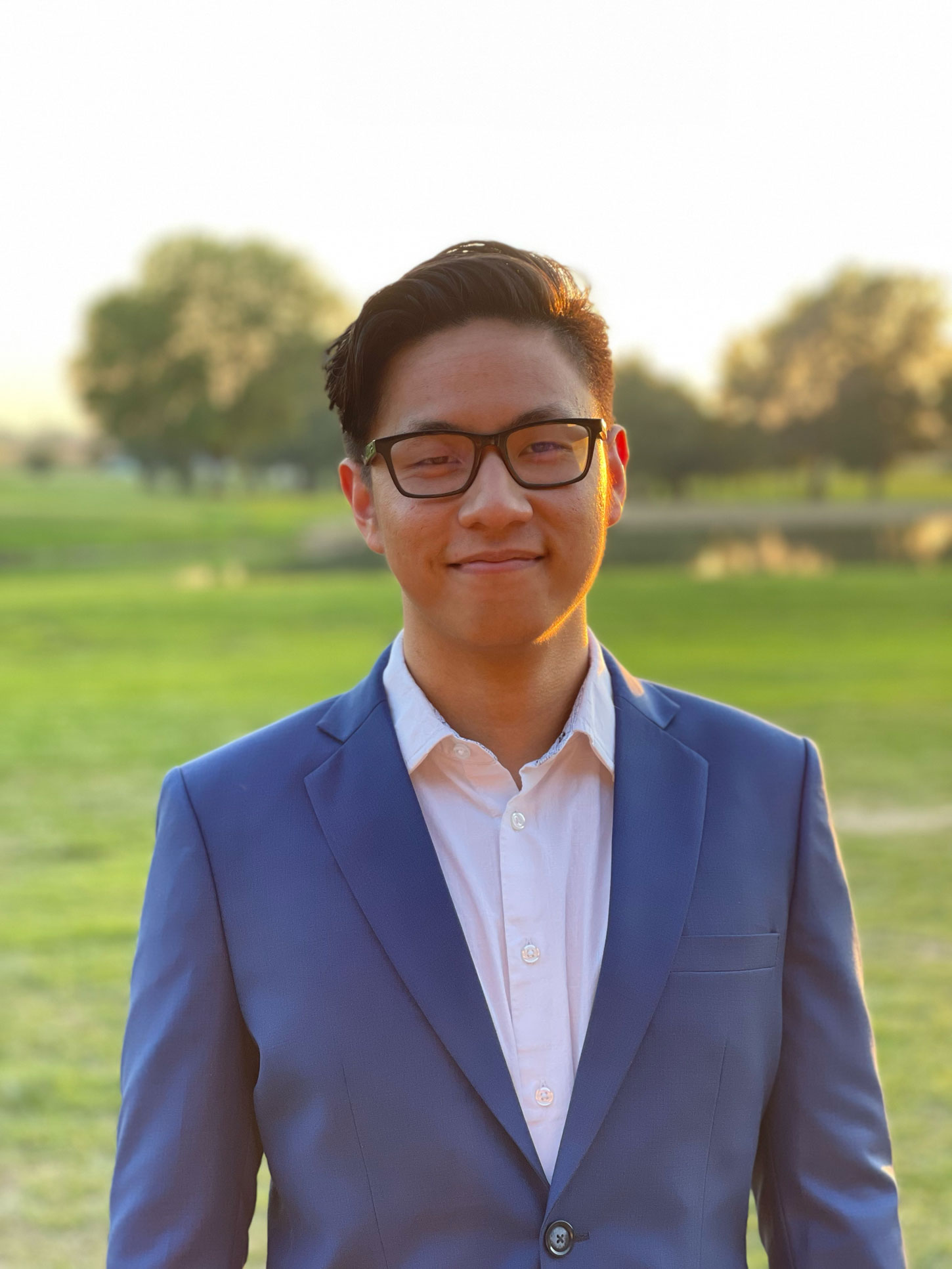 John, smiling, wearing glasses and a blue blazer outdoors at sunset